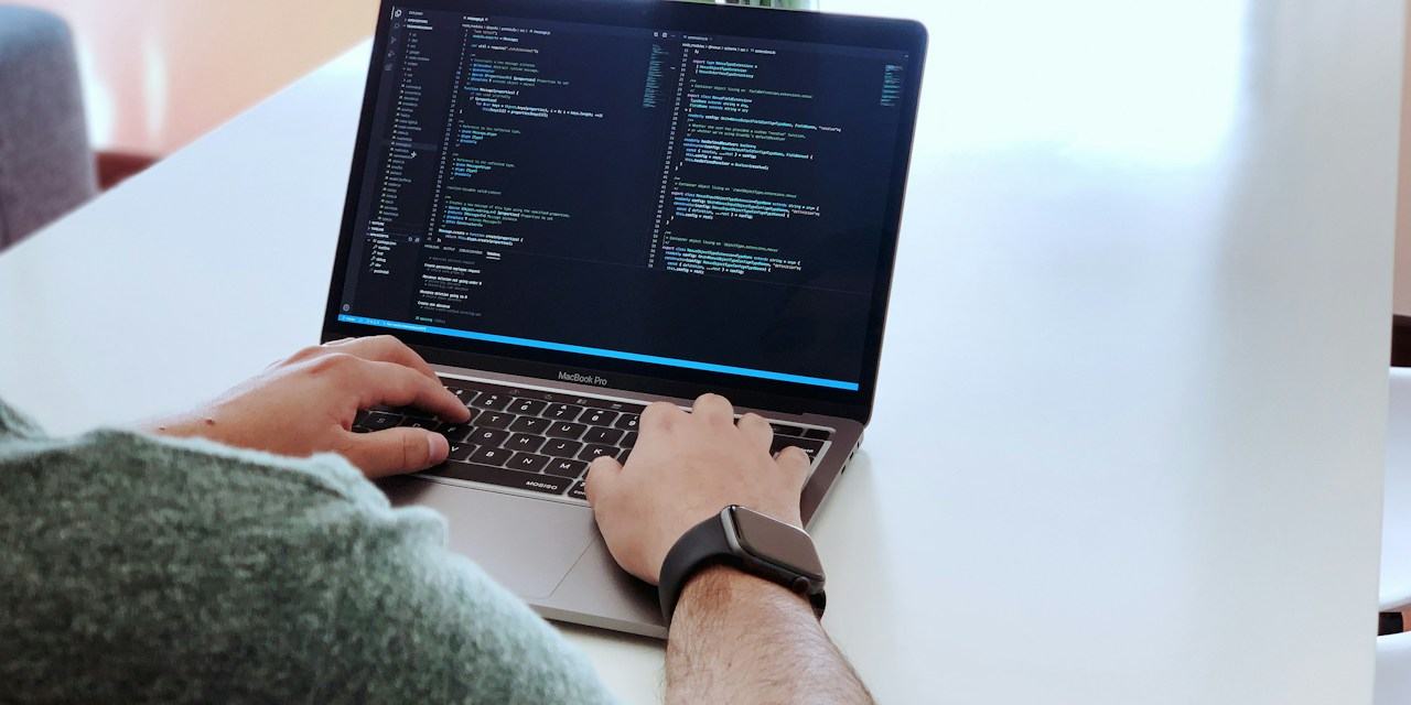 Iemand werkt aan code op een laptop aan een witte tafel.